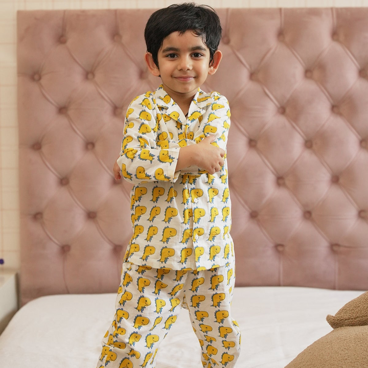 Dino Print Night Suit Set A child standing in a bedroom wearing a long sleeve night suit with dinosaur print.