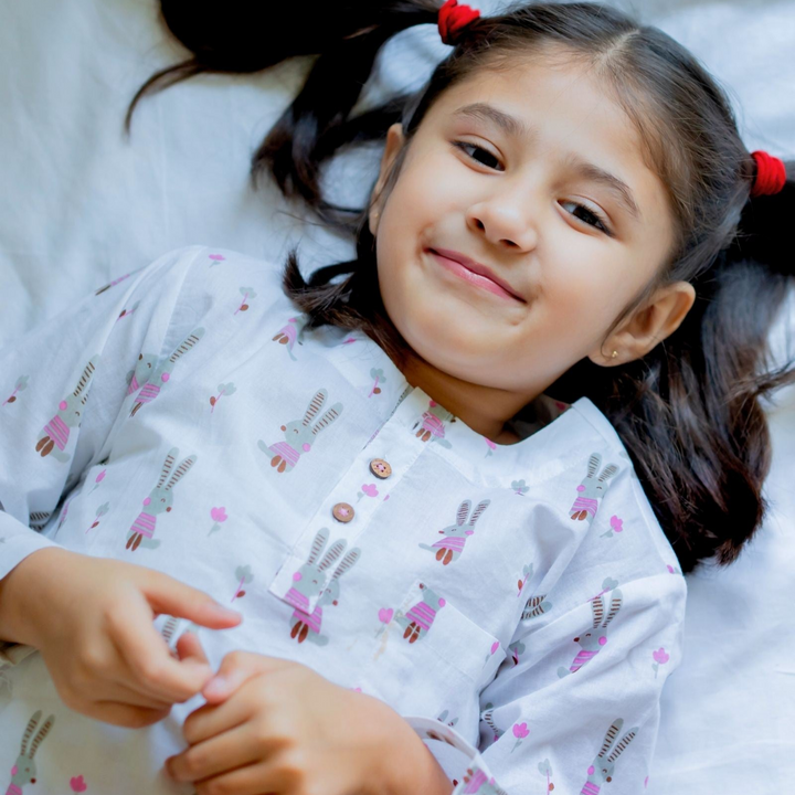 Judy Night Suit Set A girl lying on a bed wearing a white night suit with pink floral patterns and buttons.