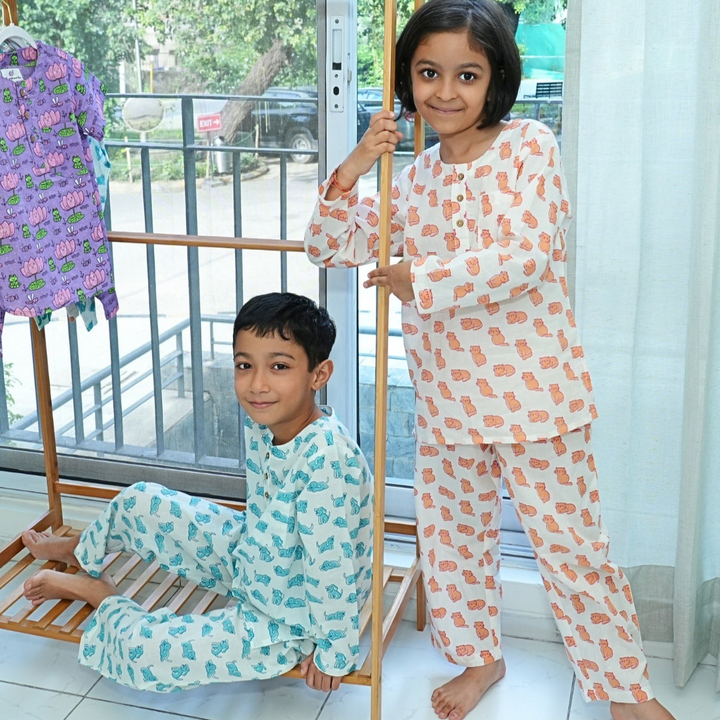 Meow Mornings PJ set night suit Two children in patterned pajamas standing next to a wooden rack indoors.