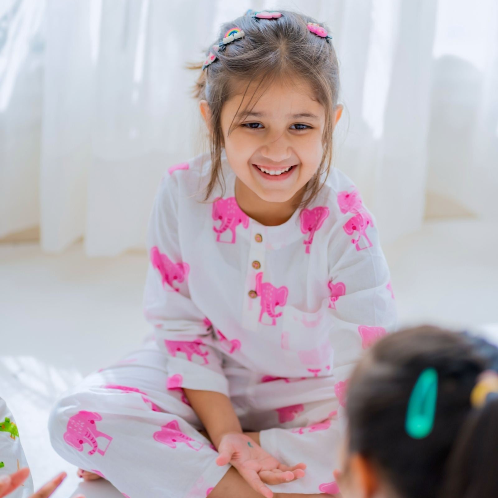 Jumbo Girl Night Suit Set A young girl smiling and sitting on a bed wearing a long sleeve night suit with a round neckline, featuring pink elephant prints on a gray background.