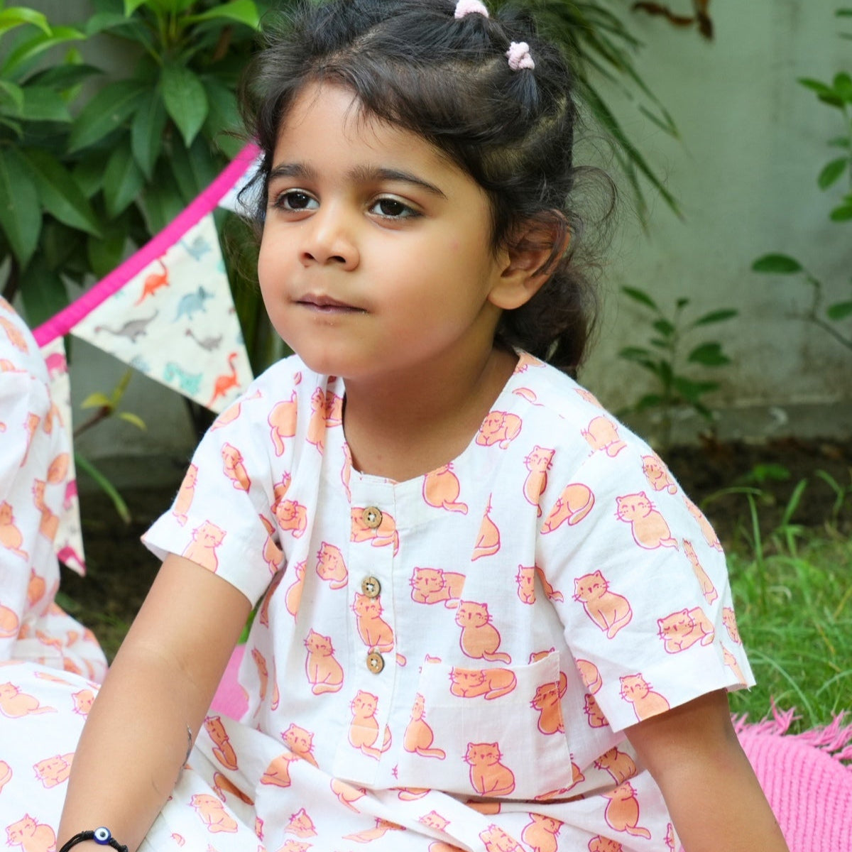 Meow Mornings PJ set night suit
Young girl in a white outfit with pink patterns sitting on grass near a small tent.