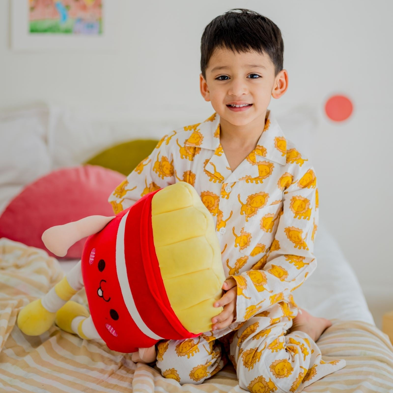 Mufasa Print Night Suit Set A child wearing a yellow and patterned night suit set with a character print, holding a stuffed toy, and sitting on a bed with a colorful cushion.