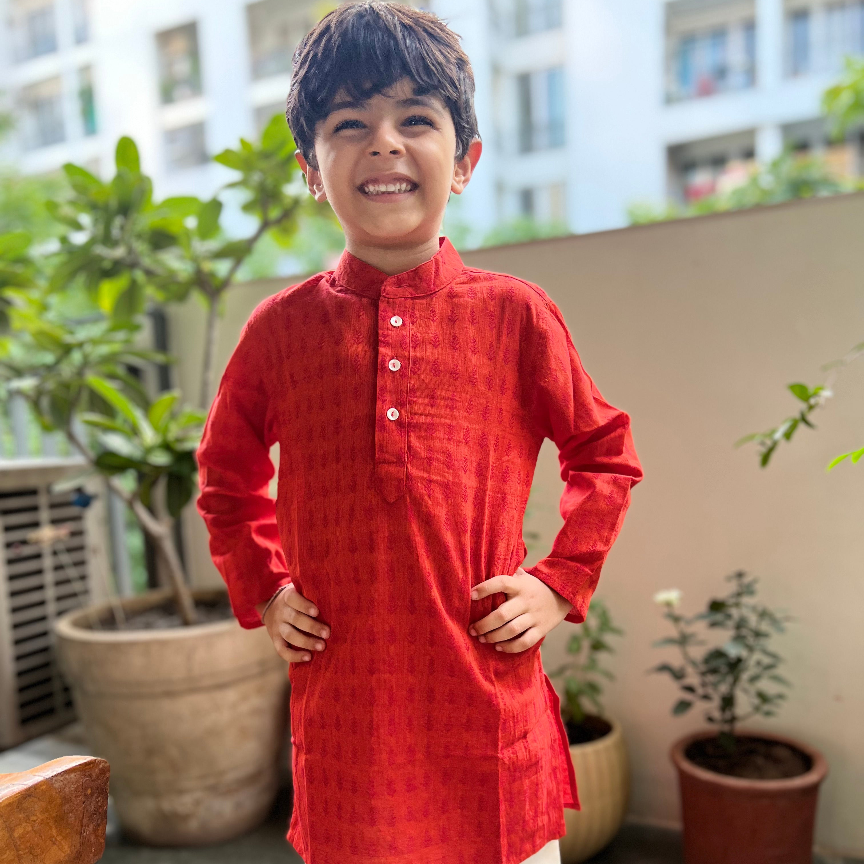 Orange Cotton Dobby Booty Kurta A child wearing a red kurta with a dobby pattern, featuring a Chinese collar, full sleeves, and front button placket, standing on a balcony with potted plants in the background.
