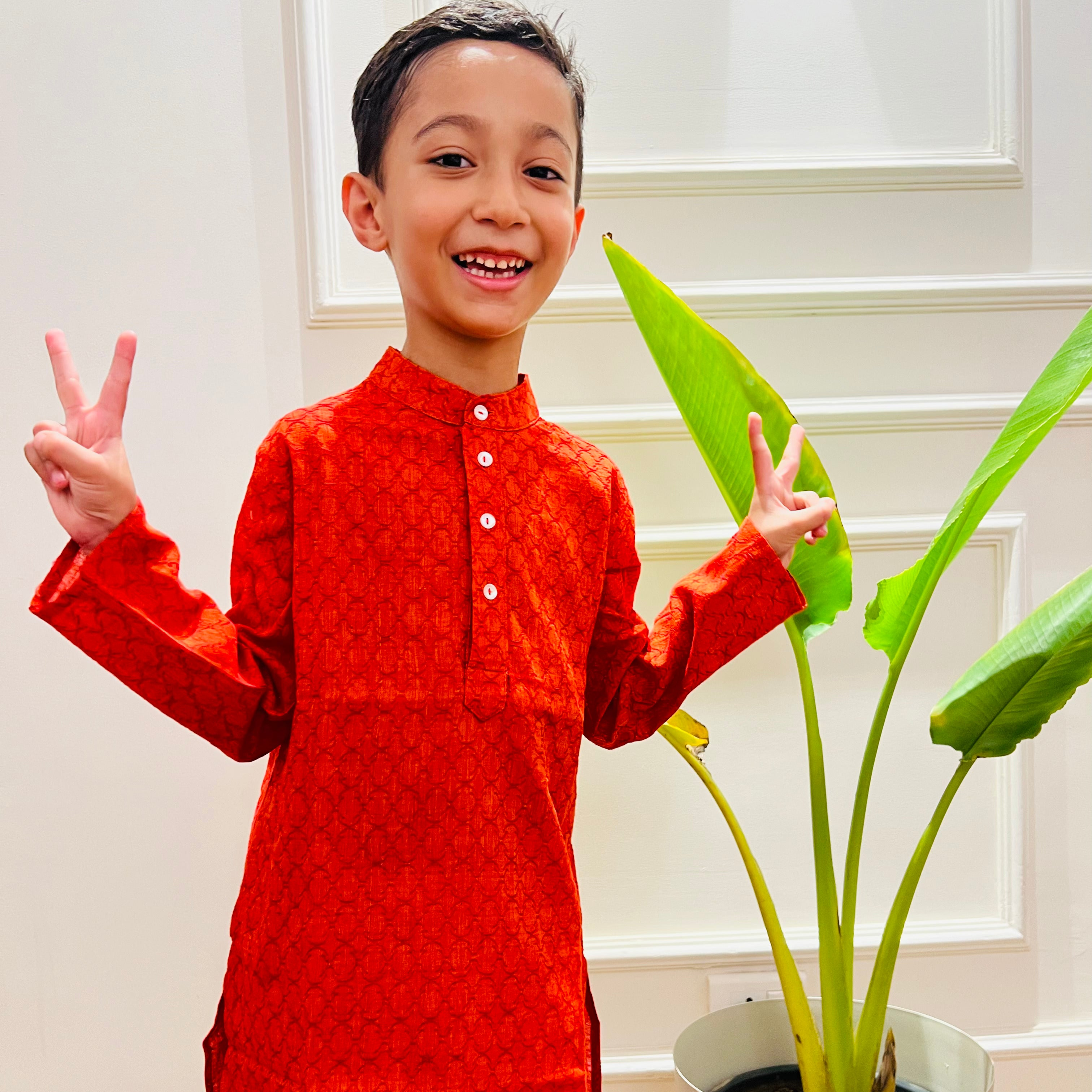 Orange Cotton Dobby Jaal Kurta A child wearing an orange long kurta with a dobby pattern, featuring side slits and a Chinese collar, standing next to a potted plant.