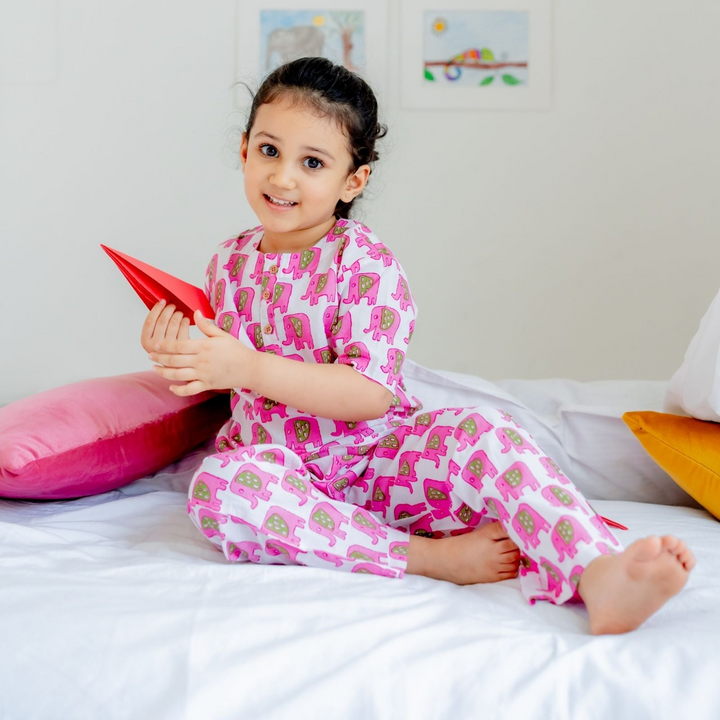 Pink Elephant Girls Night Suit Set A young girl sitting on a bed wearing a pink elephant print night suit set, holding a red book, with pillows arranged around her.