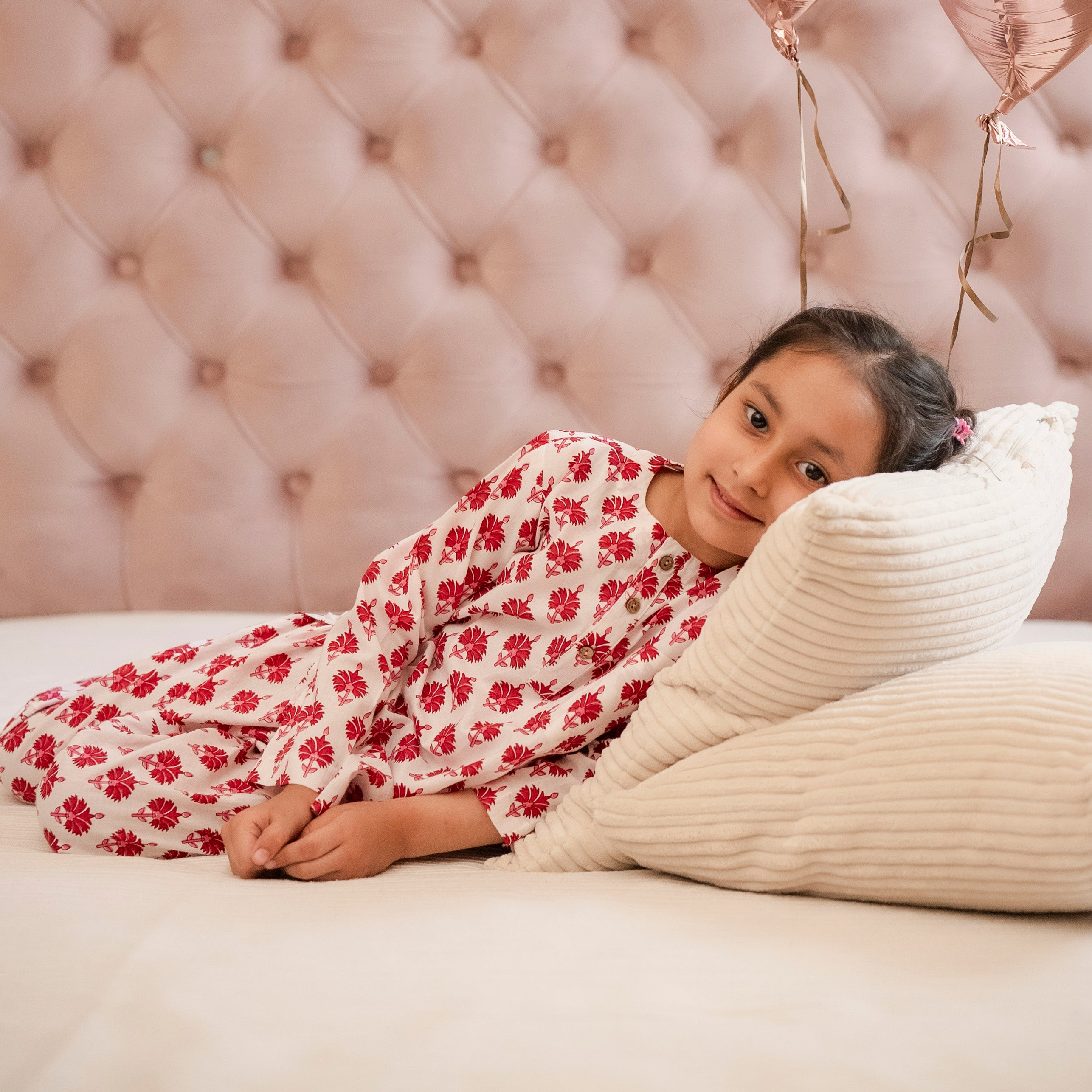 Pink Floral Night Suit Set A child lying on a bed wearing a pink floral night suit set with buttons on the front and full sleeves. The child is looking towards the camera.