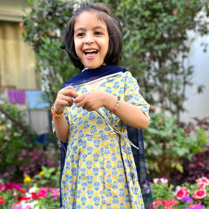 Rosa Angrakha Suit Set A young girl wearing a blue and yellow floral print angrakha suit set with a dupatta and palazzo pants, standing outdoors.