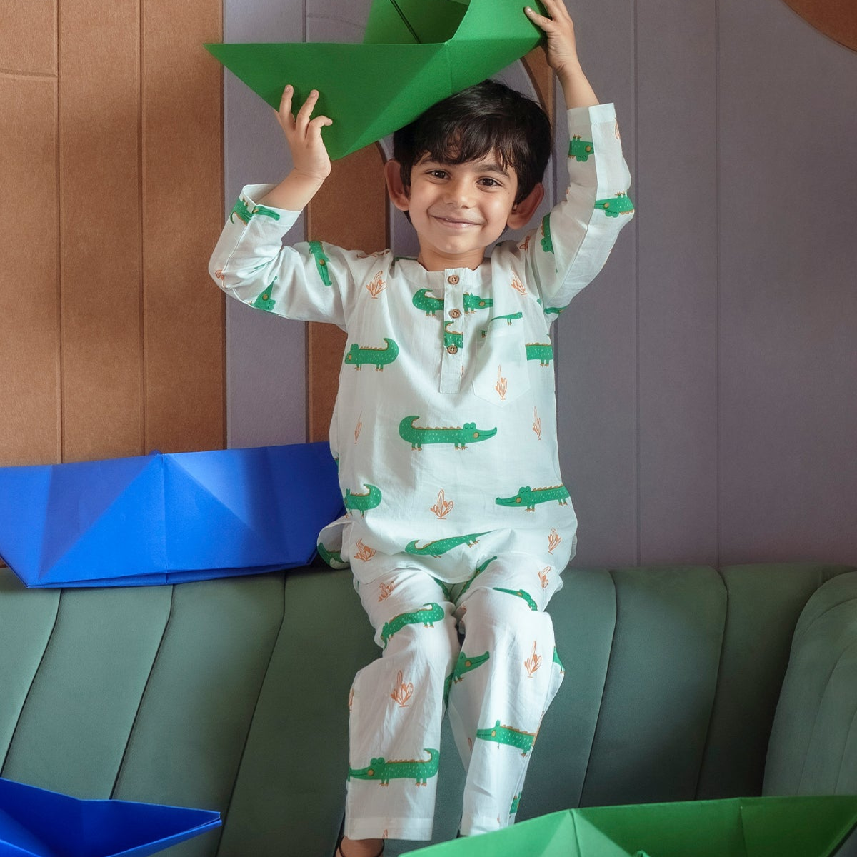 Tick Tock Night Suit Set A child wearing a long-sleeved night suit set with a pattern of clocks and stars, standing on a sofa with green tones.
